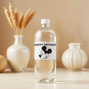 May include: A clear plastic bottle with a white cap and a label that reads "HAPPY BIRTHDAY" with a Mickey Mouse graphic. The bottle is filled with a clear liquid and is surrounded by decorative vases and dried wheat stalks.