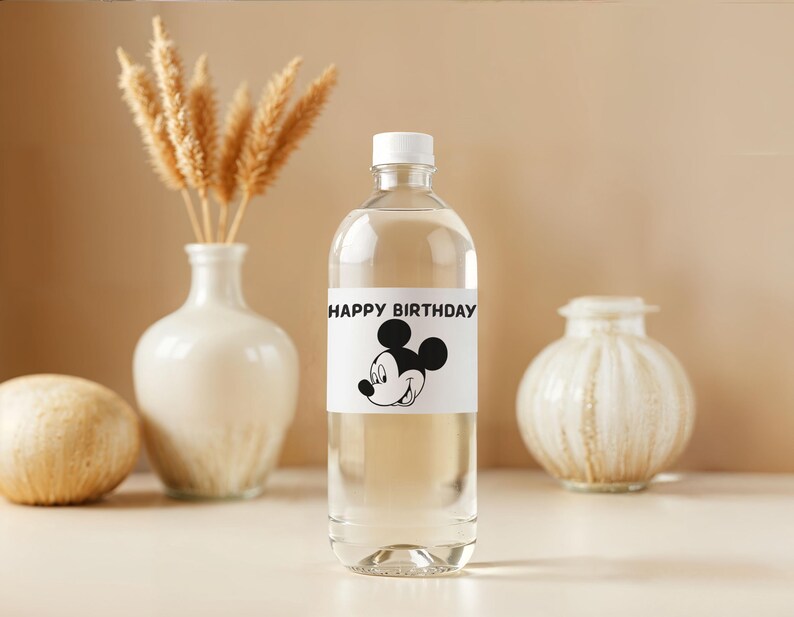 May include: A clear plastic bottle with a white cap and a label that reads "HAPPY BIRTHDAY" with a Mickey Mouse graphic. The bottle is filled with a clear liquid and is surrounded by decorative vases and dried wheat stalks.