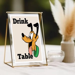 Könnte beinhalten: Ein gerahmtes Schild mit den Worten "Drink Table" und einer Cartoon-Hunde-Illustration. Das Schild befindet sich in einem goldfarbenen Metallrahmen, und der Hintergrund ist ein verschwommenes Bild einer Vase mit getrockneten Blumen und einer kleinen Glasschale.