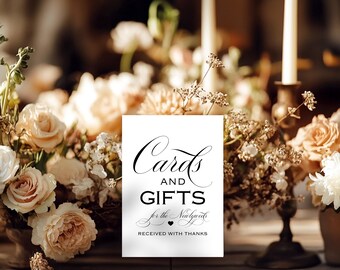 May include: A white sign with black calligraphy that reads "Cards and Gifts for the Newlyweds Received with Thanks." The sign is displayed on a metal stand, surrounded by floral arrangements and candles, creating an elegant wedding theme.