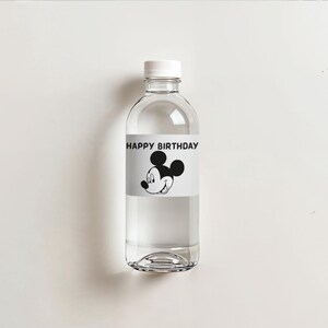 May include: A clear plastic water bottle with a white cap. The bottle has a label with the text "HAPPY BIRTHDAY" and a black and white image of Mickey Mouse. The bottle is filled with clear liquid and sits on a white surface.