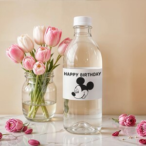 May include: A clear glass bottle with a white cap and a label that says "HAPPY BIRTHDAY" with a Mickey Mouse graphic. A bouquet of pink and white tulips sits in a glass jar. Pink roses and petals are scattered on the surface.