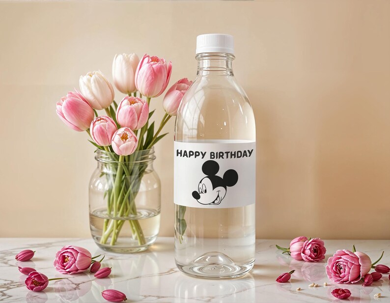 May include: A clear glass bottle with a white cap and a label that says "HAPPY BIRTHDAY" with a Mickey Mouse graphic. A bouquet of pink and white tulips sits in a glass jar. Pink roses and petals are scattered on the surface.