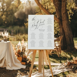 May include: A white wedding seating chart on a wooden easel, with the words "The Best Day Ever" in elegant script. The names of the wedding guests are listed below. The sign is displayed outdoors in a natural setting.