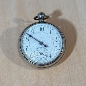 May include: A silver pocket watch with a white face and black numerals. The watch has a small seconds dial and a winding stem at the top. The hands are black and the watch is set against a light brown wooden surface.