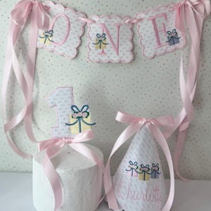May include: A pink and white "ONE" banner with a matching cake topper and party hat. The banner and topper feature embroidered gift box designs. The party hat is personalized with the name "Charlotte". All items are decorated with pink ribbons.