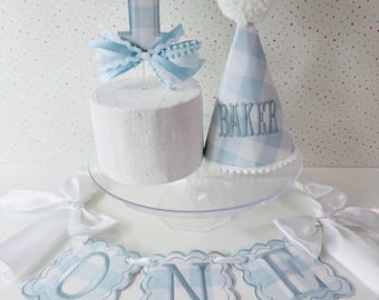 May include: A light blue and white gingham party set featuring a cake topper with the number one, a cone-shaped party hat embroidered with the name BAKER and topped with a white pom-pom, and a banner spelling out the word ONE with scalloped edges.