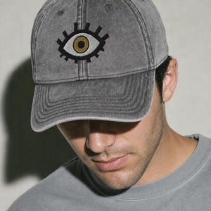 May include: A charcoal gray baseball cap with a distressed, washed-out finish. The front panel features an embroidered graphic of a stylized eye with a gold iris, a white sclera, and thick black outlines with radiating lashes.