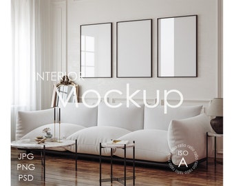 May include: A white sofa with three pillows sits in a living room with three black framed pictures on the wall. The sofa is in front of a large window with white curtains. There are two small tables in front of the sofa, one with a vase of flowers and the other with a book. The floor is made of wood.