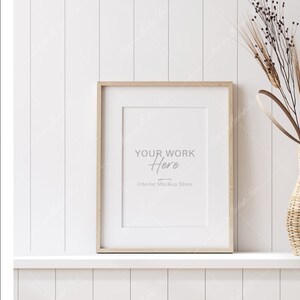 Peut inclure: Un cadre en bois clair avec un passe-partout blanc et un fond blanc. Le cadre est posé sur une étagère blanche avec un mur blanc derrière. Le texte "YOUR WORK Here Interior Mockup Store" est imprimé sur le fond blanc. Un vase avec des fleurs séchées est sur le côté droit de l'image.