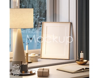 May include: A white picture frame with a blank canvas sits on a wooden table next to a lit candle and a lamp with a beige lampshade. The table is in front of a window with a view of a snowy forest. The text "INTERIOR MOCKUP" is visible in the image.