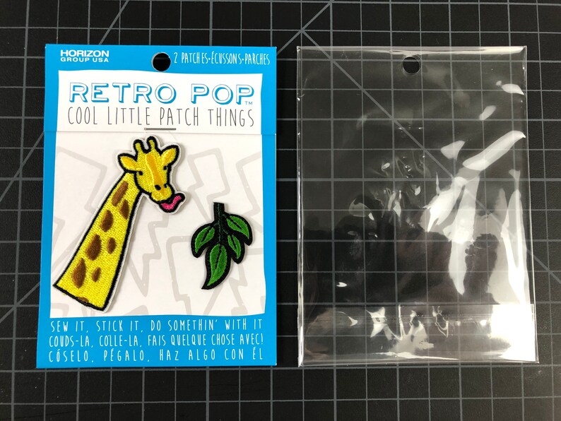 以下が含まれることがあります： アイロン接着式のワッペン2枚。1枚は葉っぱを食べているキリン、もう1枚は緑の葉っぱ。ワッペンは白いカードに貼られており、黒い文字で「レトロポップクールリトルパッチシングス」と「縫う、貼る、何かする」と書かれている。