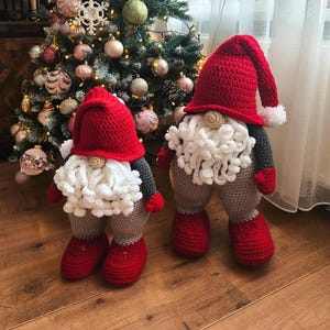 May include: Two handmade crochet gnomes with red hats, white beards, and grey and red clothing. The gnomes stand before a decorated Christmas tree with baubles and lights. The Passionate Crafter logo is visible in the background.