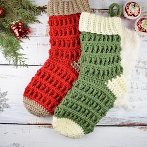 May include: Two crocheted Christmas stockings, one red and one green, with white and brown accents. The stockings are hanging on a white wooden background.