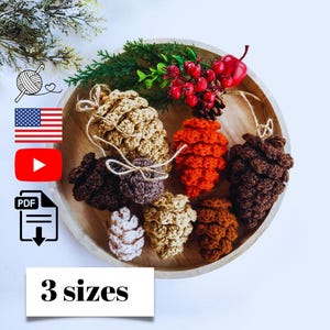 May include: A collection of handmade crochet pine cones in various shades of brown, orange, and white, arranged on a wooden plate with greenery and red berries. The image includes the text "3 sizes".