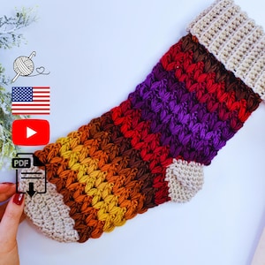 May include: A handmade crochet Christmas stocking in a gradient of colors, from beige to orange, red, and purple. The stocking features a textured stitch pattern and a beige cuff and toe. The image includes a PDF icon, a YouTube icon, and the American flag.