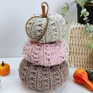 Op de afbeelding: Een stapel van drie gehaakte pompoenen in neutrale kleuren. De bovenste pompoen is beige, de middelste is roze en de onderste is bruin. Elke pompoen heeft een textuurpatroon en een bruine steel. Op de achtergrond een kleine oranje kalebas en een gevlochten mand.