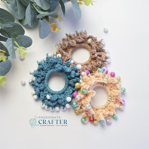 May include: Three handmade crochet hair scrunchies in teal, tan, and peach, each adorned with decorative beads. The scrunchies are arranged on a white surface with eucalyptus leaves and the text "PASSIONATE CRAFTER".