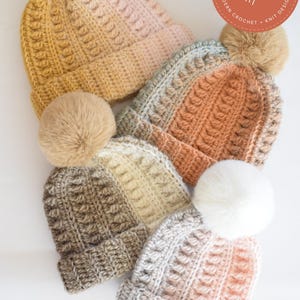 May include: A collection of handmade crochet beanies in various colors, including mustard yellow, peach, and gray. Each hat features a textured stitch pattern and a fluffy pom-pom on top. The beanies are arranged on a white surface.