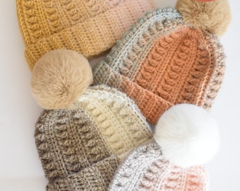 May include: A collection of handmade crochet beanies in various colors, including mustard yellow, peach, and gray. Each hat features a textured stitch pattern and a fluffy pom-pom on top. The beanies are arranged on a white surface.