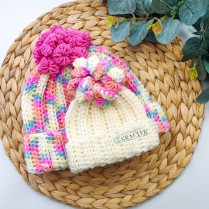 May include: Two crocheted hats. One is ivory with a floral accent and the text "PASSIONATE CRAFTER". The other is multi-colored with pink, yellow, and blue hues, and a large pink floral accent. Both hats are on a woven mat.