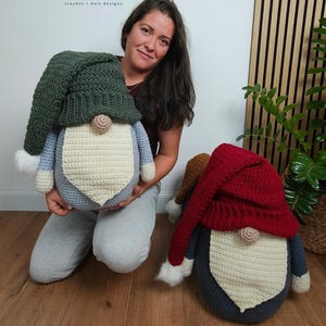 May include: Two large handmade crochet gnomes featuring pointed hats, round noses, and long beards. One gnome has a dark green hat and light blue body, while the other has a red hat and dark blue body. Text reads PASSIONATE CRAFTER crochet knit designs.