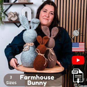 Może przedstawiać: Trzy szydełkowe figurki króliczków w odcieniach niebieskiego, brązowego i beżowego, prezentowane na drewnianej tacy. Każdy króliczek ma długie uszy i jest związany sznurkiem. Widoczny jest tekst "3 Sizes" i "Farmhouse Bunny".