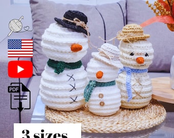 May include: Three crocheted snowmen of varying sizes, each with a unique hat, scarf, and carrot nose. The snowmen are white with black stitched eyes and mouth details. One snowman has a black hat, another a straw hat, and the third a white hat.