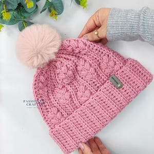 May include: A dusty pink crocheted beanie featuring a textured cable knit pattern and a large, matching faux fur pom-pom on top. The hat has a wide ribbed brim and a small rectangular metal tag labeled Handmade. The text PASSIONATE CRAFTER is visible.