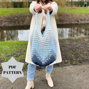 May include: A crocheted tote bag with a gradient of blue colours. The bag has a wide, open top and is made with a simple stitch pattern. The bag is being held by a person wearing a white coat and blue jeans.