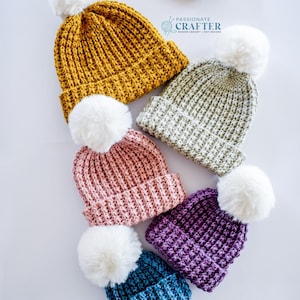 May include: A collection of six handmade knit beanies in various colors, including mustard yellow, sage green, pink, purple, and blue. Each beanie features a ribbed design and a large white pom-pom on top. The beanies are arranged on a white surface.