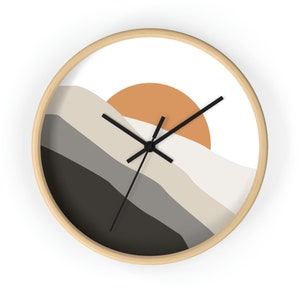 May include: A round wooden wall clock with a white face and black hands. The clock face features a stylized abstract design of a sunset over hills in shades of gray, beige, and orange.