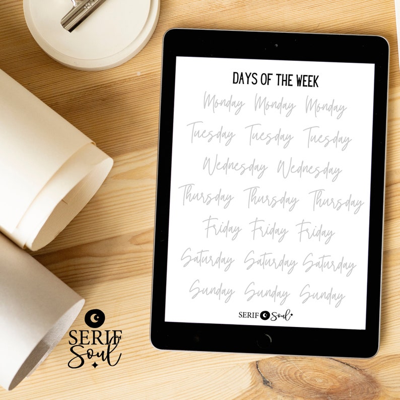 Puede incluir: Una tableta digital muestra el texto "DAYS OF THE WEEK" en una fuente manuscrita. Los d&iacute;as de la semana se enumeran varias veces. La tableta est&aacute; sobre una superficie de madera, con papel enrollado y un objeto circular blanco en el fondo.