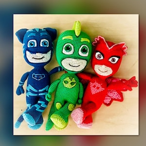 Puede incluir: Tres muñecos de ganchillo hechos a mano al estilo de los personajes de PJ Masks. Los muñecos son azules, verdes y rojos, con caras sonrientes y trajes detallados. El muñeco azul tiene rasgos de gato, el verde un diseño de gecko y el rojo una apariencia de murciélago.