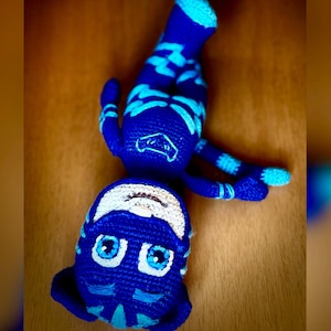 May include: A blue crocheted stuffed toy with a tiger face, white eyes, and a blue and white striped pattern. The toy is lying on a wooden surface.