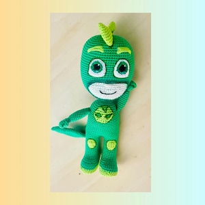 May include: Green crocheted plush toy of a superhero character with a green mask, a green suit, and a green tail. The toy has a white smile and large green eyes.