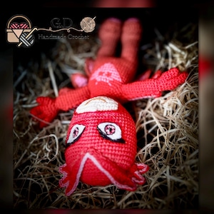 May include: Red crocheted plush toy of a superhero character with a mask and a star on the chest. The toy is lying on a bed of brown straw.