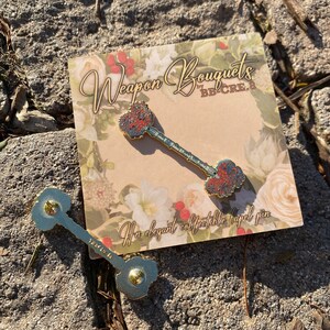 K&ouml;nnte beinhalten: Goldfarbene Anstecknadel in Form eines Schl&uuml;ssels mit roten und blauen floralen Details an Kopf und Basis. Die Nadel liegt auf einer Karte mit floralem Thema mit dem Text "Weapon Bouquets" und "An elegant collectible lapel pin."