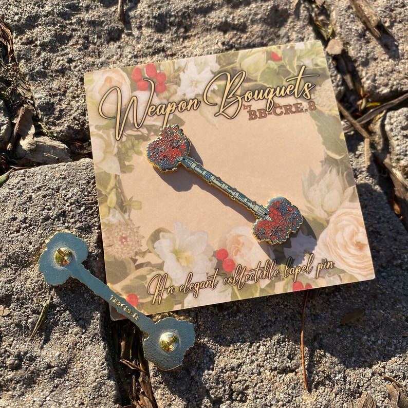 K&ouml;nnte beinhalten: Goldfarbene Anstecknadel in Form eines Schl&uuml;ssels mit roten und blauen floralen Details an Kopf und Basis. Die Nadel liegt auf einer Karte mit floralem Thema mit dem Text "Weapon Bouquets" und "An elegant collectible lapel pin."