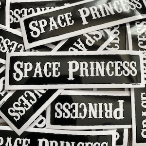 May include: A stack of black and white stickers with the text "SPACE PRINCESS" printed in a bold, white font.