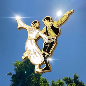 May include: A gold-colored enamel pin depicting a couple dancing. The woman is in a white dress, and the man is in a black and yellow outfit. The pin is set against a blue sky background.