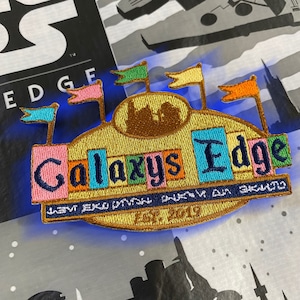 May include: A colorful embroidered patch with the text "Galaxy's Edge" in a stylized font. The patch features a brown castle-like structure with flags in various colors, including blue, green, pink, and orange.