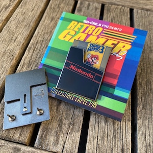 May include: A black and gold enamel pin featuring a Nintendo Entertainment System cartridge with a Super Mario Bros. 3 game insert. The pin is on a card that reads "BB-CRE.8 PRESENTS RETRO GAMER Series COLLECTIBLE LAPEL PIN".
