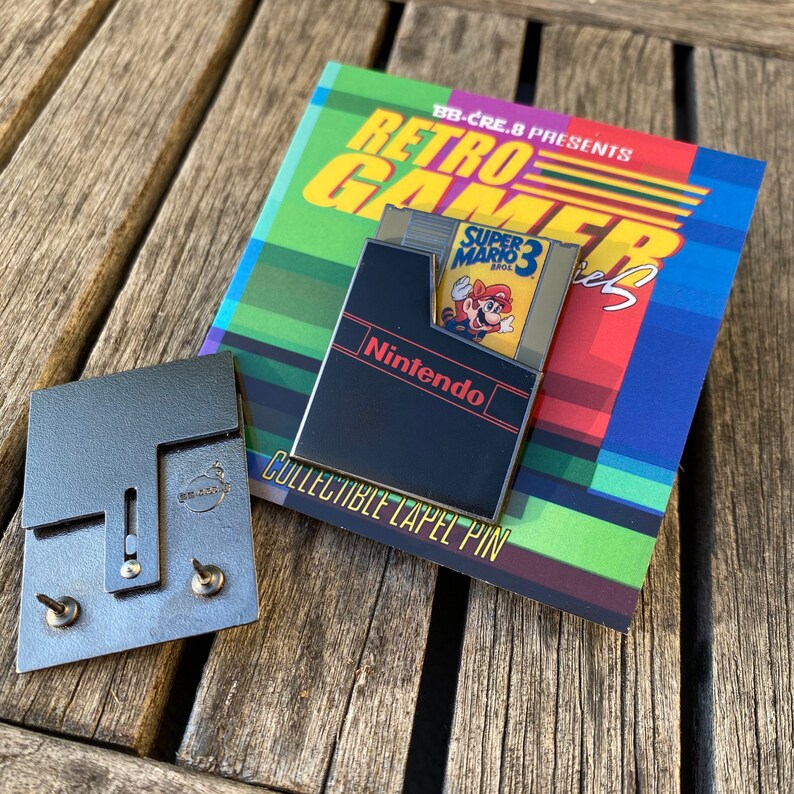 May include: A black and gold enamel pin featuring a Nintendo Entertainment System cartridge with a Super Mario Bros. 3 game insert. The pin is on a card that reads "BB-CRE.8 PRESENTS RETRO GAMER Series COLLECTIBLE LAPEL PIN".