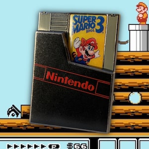 May include: A black and red Nintendo game cartridge with a silver trim. The cartridge is labelled "Super Mario Bros. 3". The cartridge is partially inserted into a black Nintendo Entertainment System game case.
