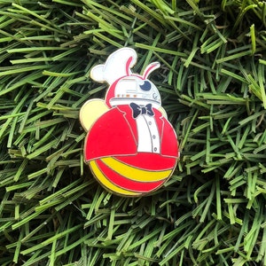 May include: A red and yellow enamel pin of a robot bee wearing a tuxedo. The bee has a white chef's hat and a black bow tie.