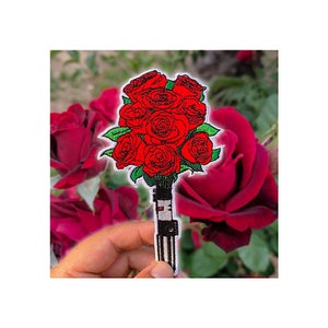 May include: Embroidered patch featuring a bouquet of red roses with green leaves. The roses are tightly clustered, with a detailed design. The patch has a black and white striped handle, and is set against a blurred background of red roses.