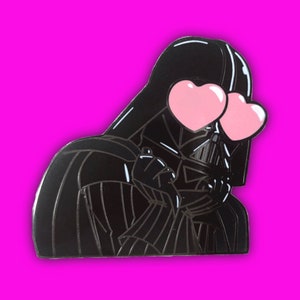 May include: A black enamel pin featuring Darth Vader with pink heart-shaped eyes. The pin is set against a bright pink background. The design is a stylized, cartoon-like representation of the Star Wars character.