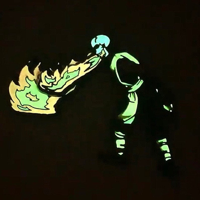May include: A green and yellow glow-in-the-dark enamel pin featuring a character with a green and yellow flame in their hand.