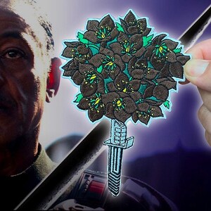May include: Embroidered patch featuring a bouquet of dark brown flowers with green and yellow accents, atop a stylized dagger. The patch has a light blue border and is held against a blurred background.